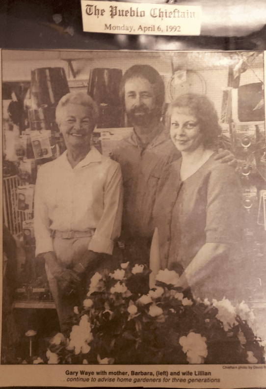 Group of three smiling people posing with flowers in front of a shop. The Pueblo Chieftain newspaper header is visible.