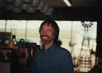 Man smiles inside a store, wearing blue shirt. Background includes gardening items, window, and a small windmill.