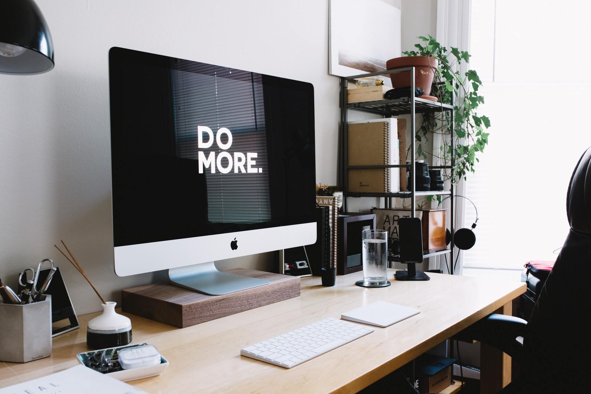 Modern desk setup with iMac displaying “DO MORE.” and shelves with plants and office supplies