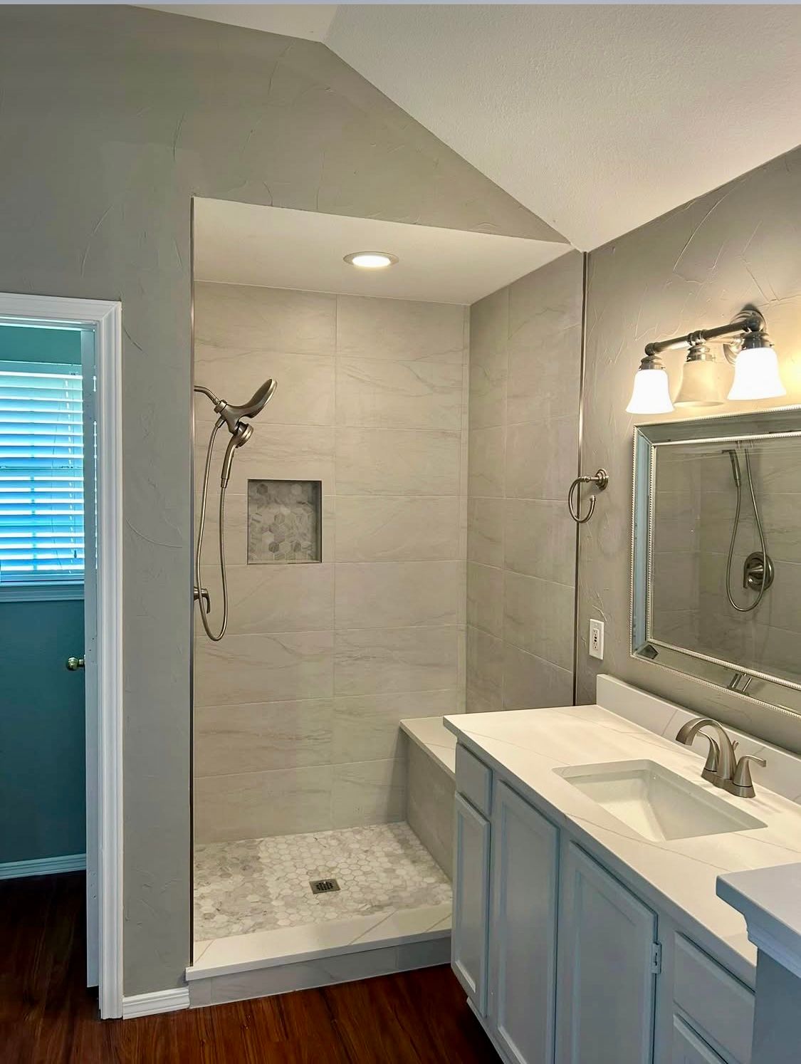 Modern bathroom with a white vanity, a large shower with a built-in bench, and gray tiled walls. A window is partially visible.