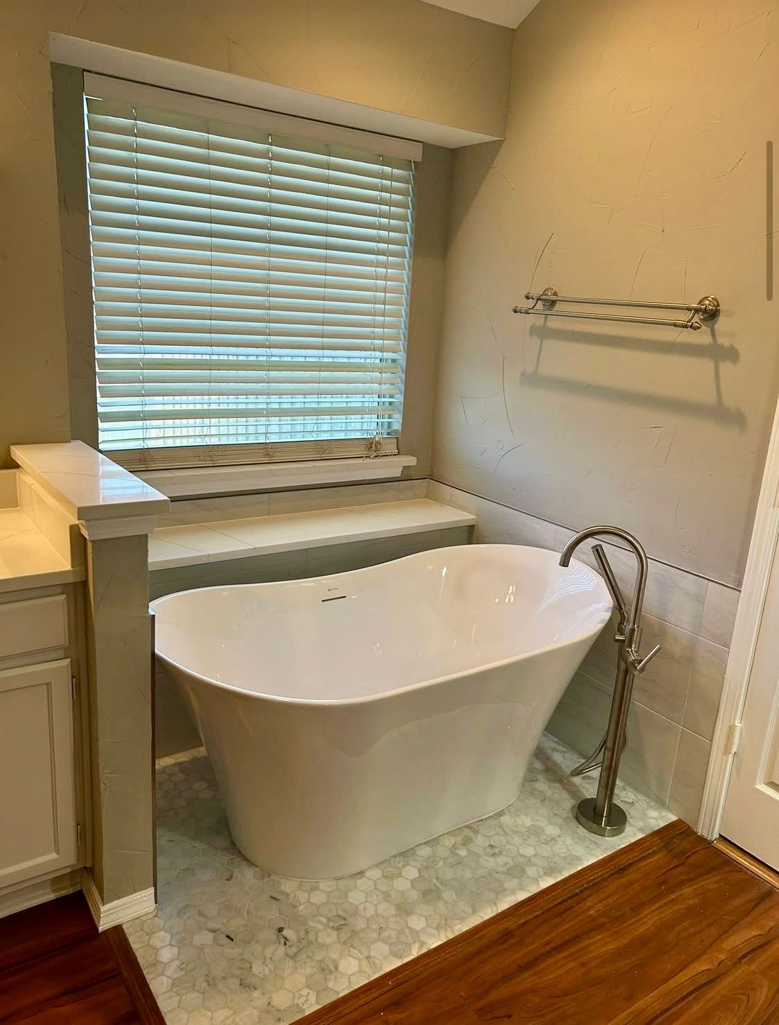 A white freestanding bathtub sits under a window with blinds. The bathroom has tan walls, wood floors, and a towel rack.