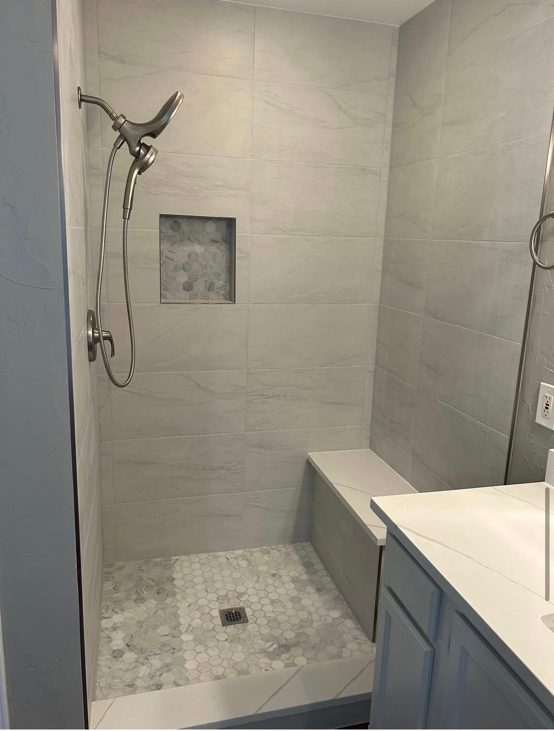A remodeled bathroom shower with light gray tiles and a built-in bench. A niche holds supplies, and the shower head is visible.