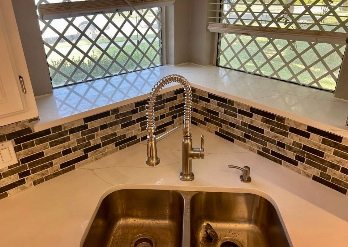 Stainless steel kitchen sink with a pull-down faucet, on a white countertop with a mosaic tile backsplash.