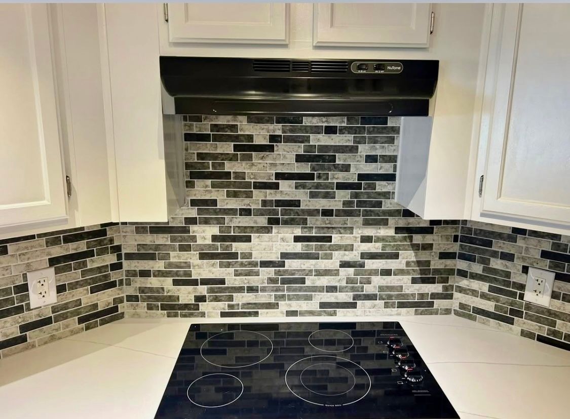 Kitchen backsplash with black, gray, and white mosaic tile above a stovetop and white countertops.