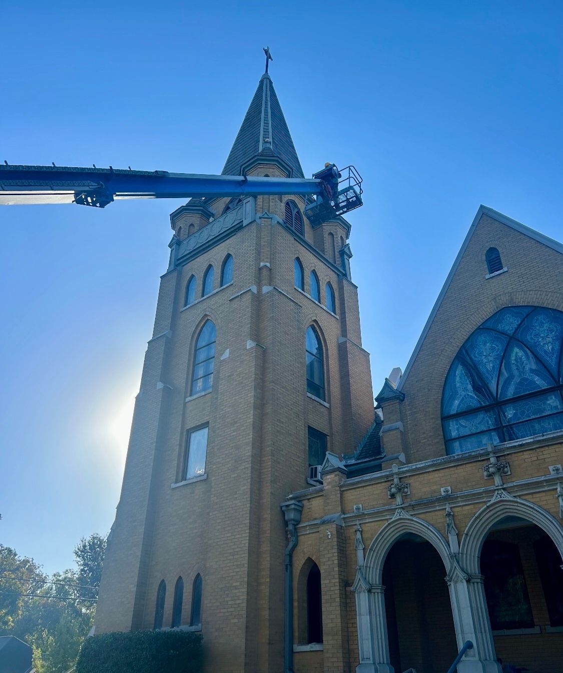 Tall brick church tower with a lift basket near the top, under a blue sky with the sun shining.