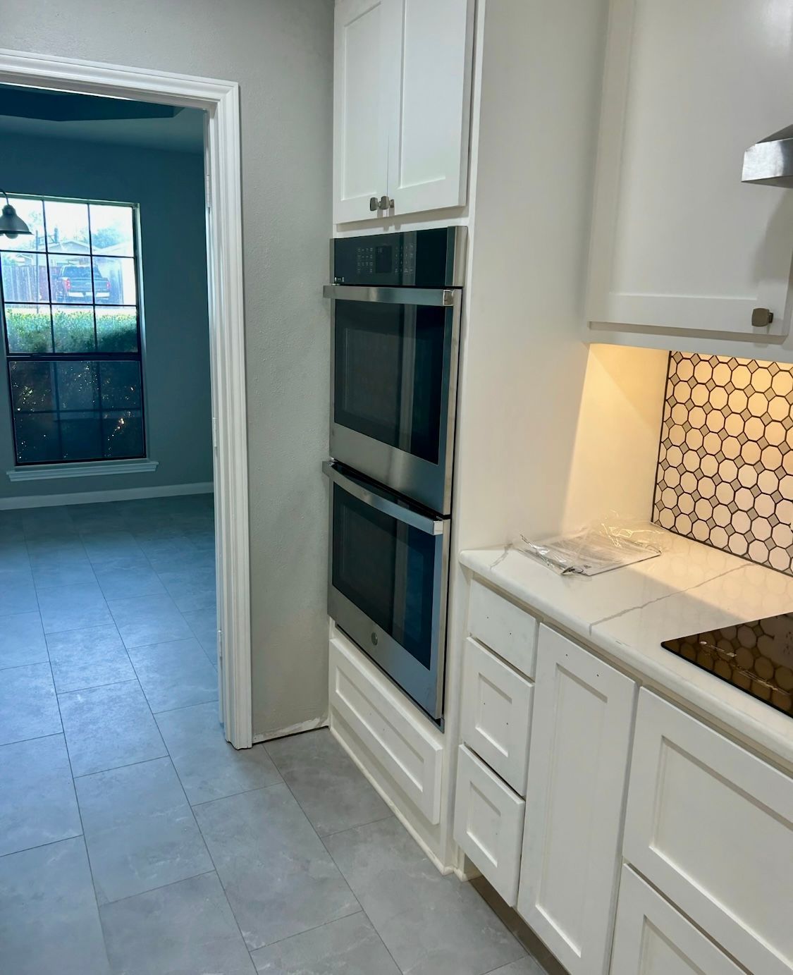 Kitchen with white cabinets, double oven, and tiled backsplash. Gray flooring and view of a room.
