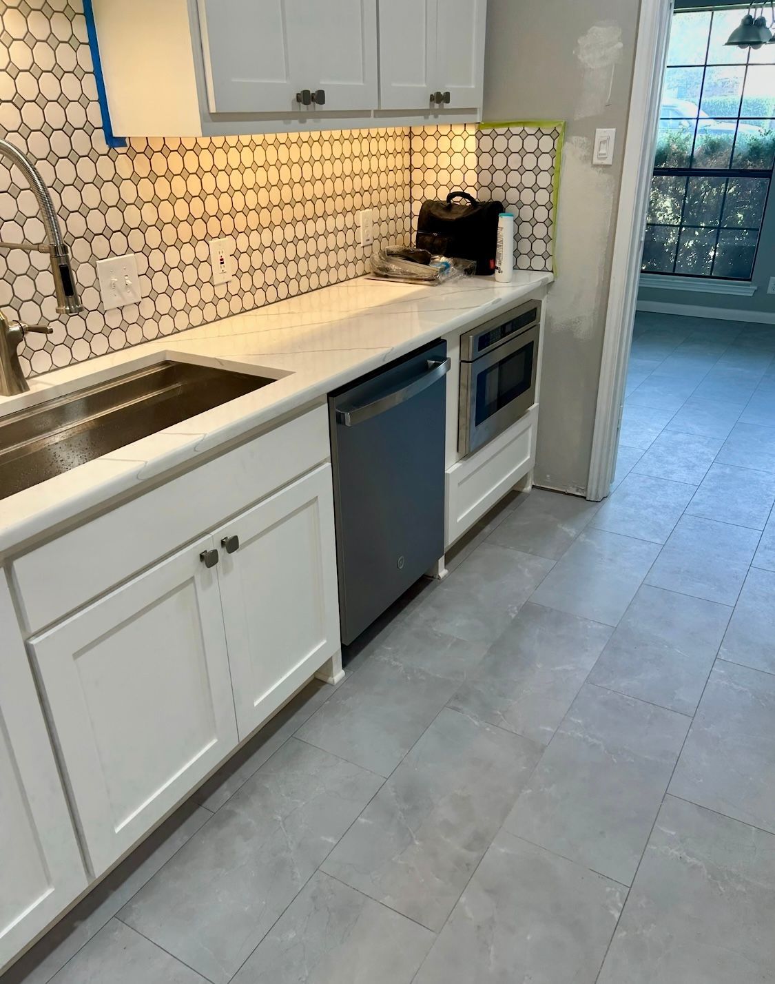White kitchen with sink, cabinets, dishwasher, microwave. Gray floor, tiled backsplash, countertop, and a window.