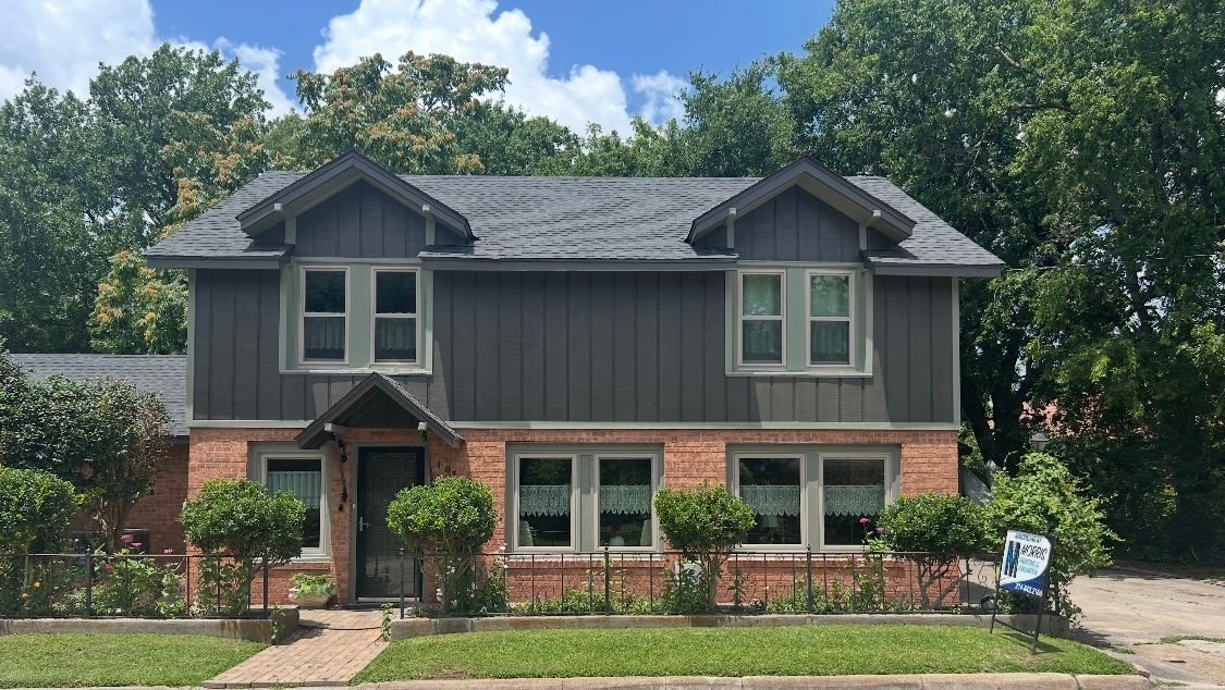 Two-story house with gray siding, brick base, dormers, windows, and a small front yard with bushes.