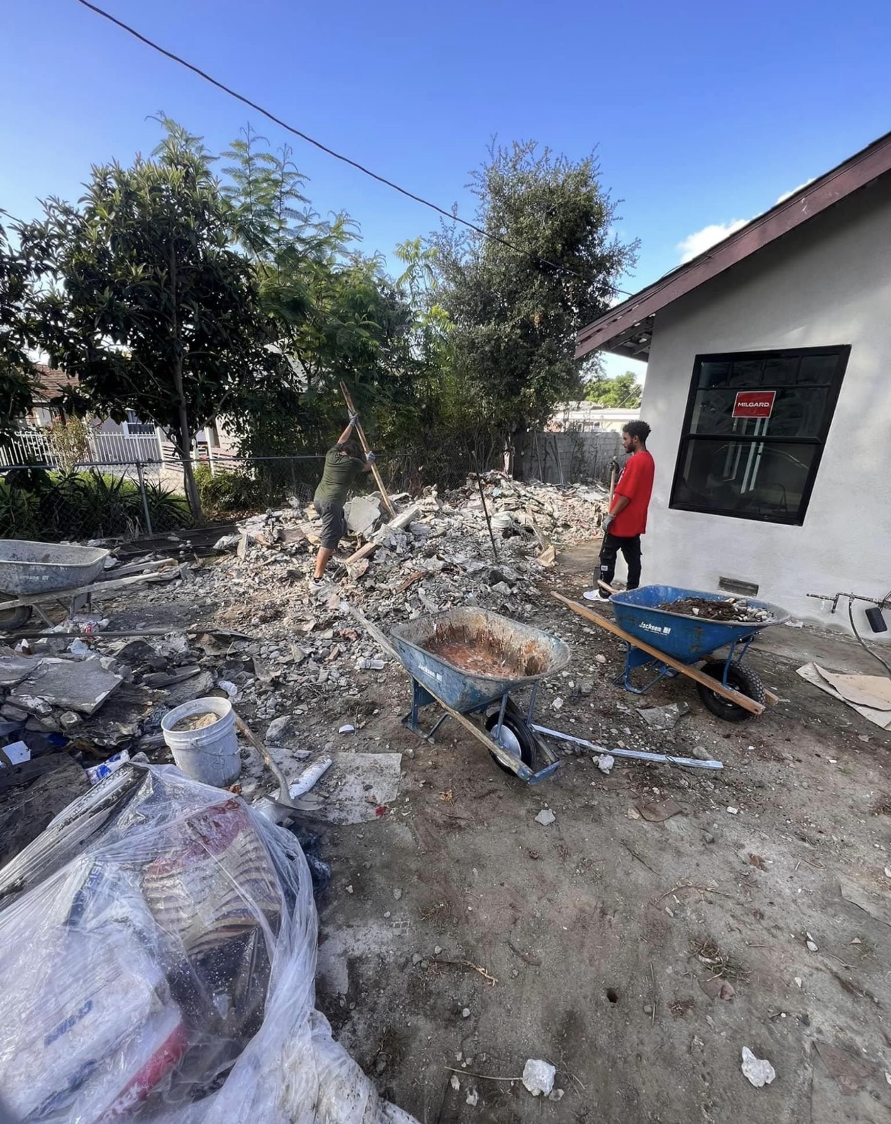 A man is standing next to a wheelbarrow in front of a house.