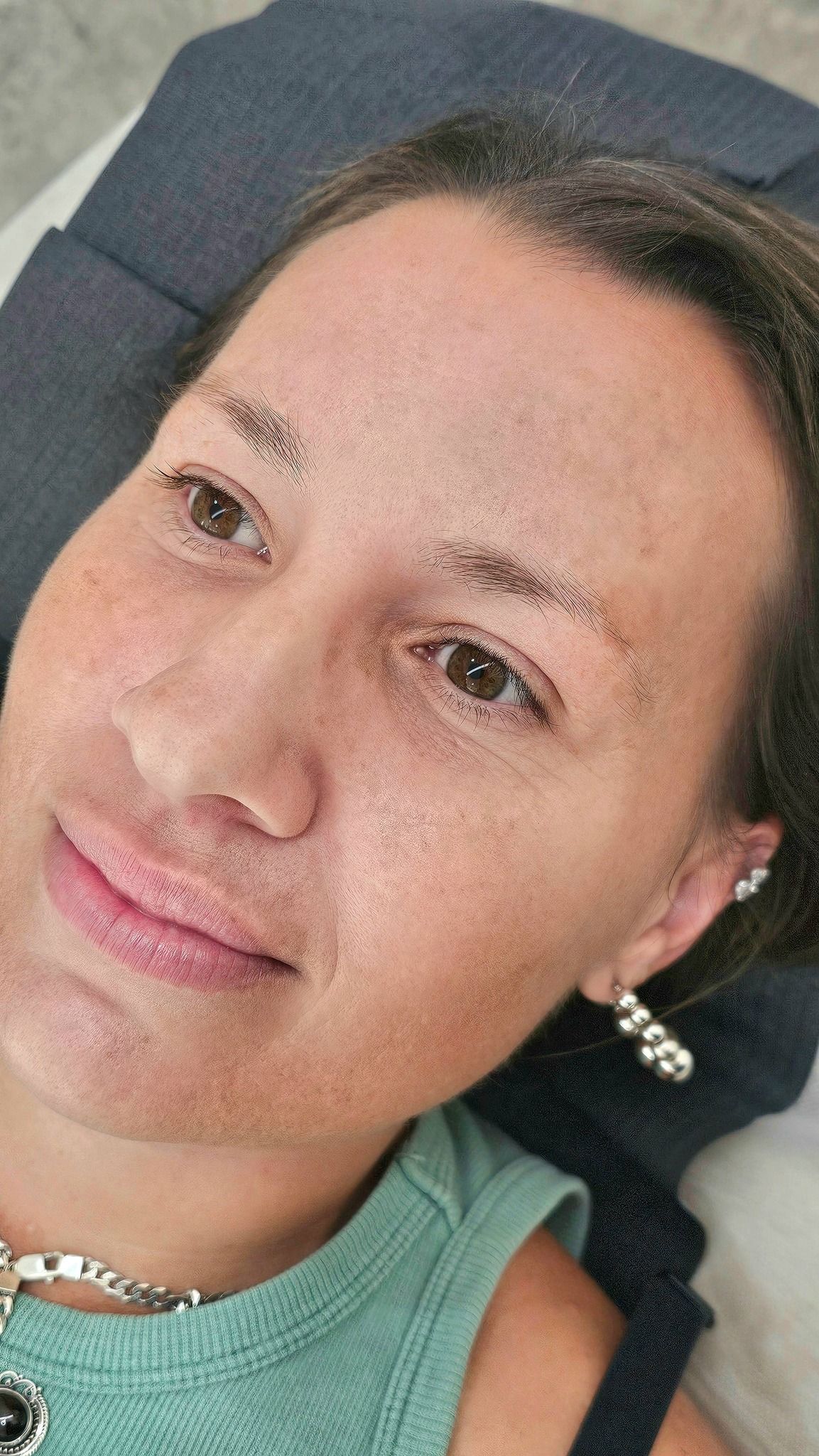 Woman with faint eyebrow tattoos, wearing earrings and a necklace, looking at the camera.