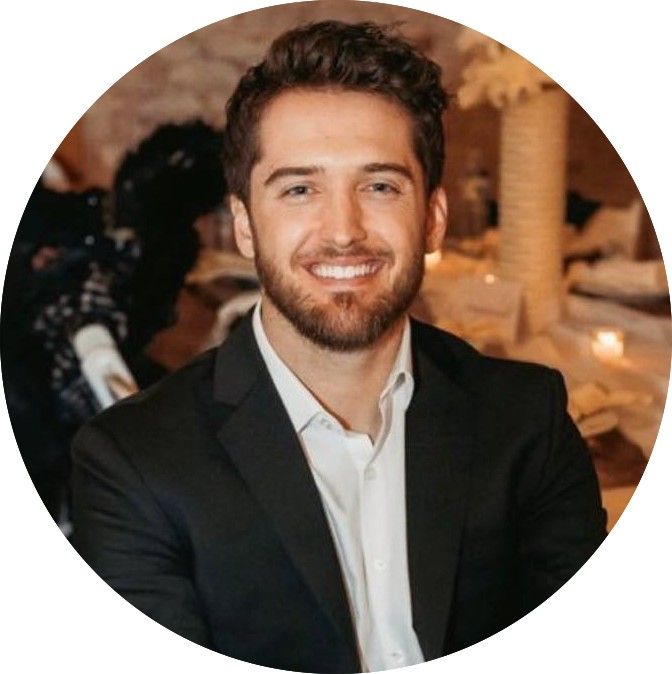 Smiling man in a black suit and white shirt, with a beard. Indoor setting, blurred background.