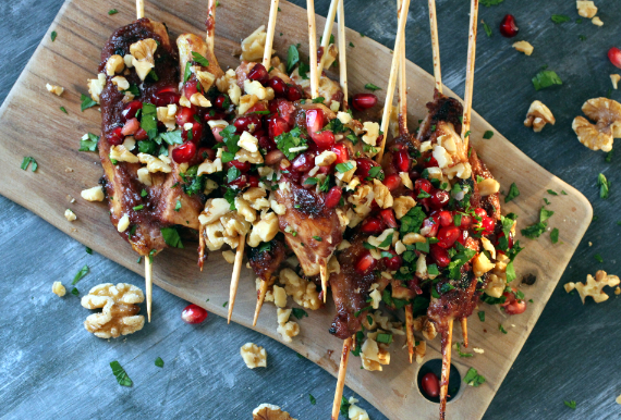 A wooden cutting board topped with skewers of food with pomegranate seeds and walnuts.