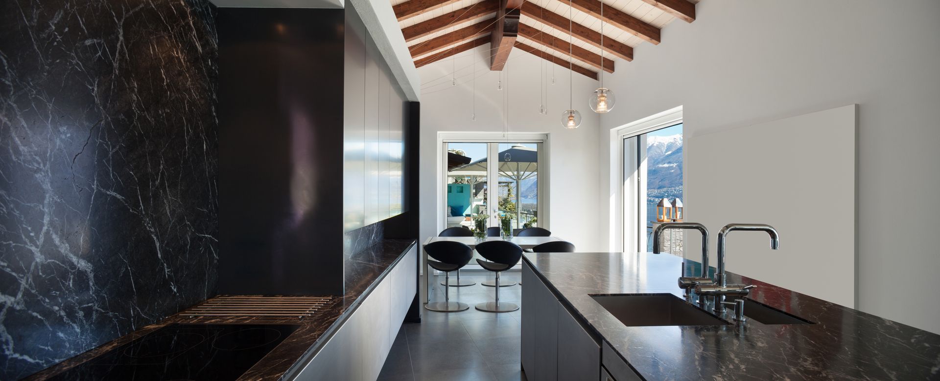 Modern kitchen with dark marble countertops, exposed wood beams, and a bright view through glass doors