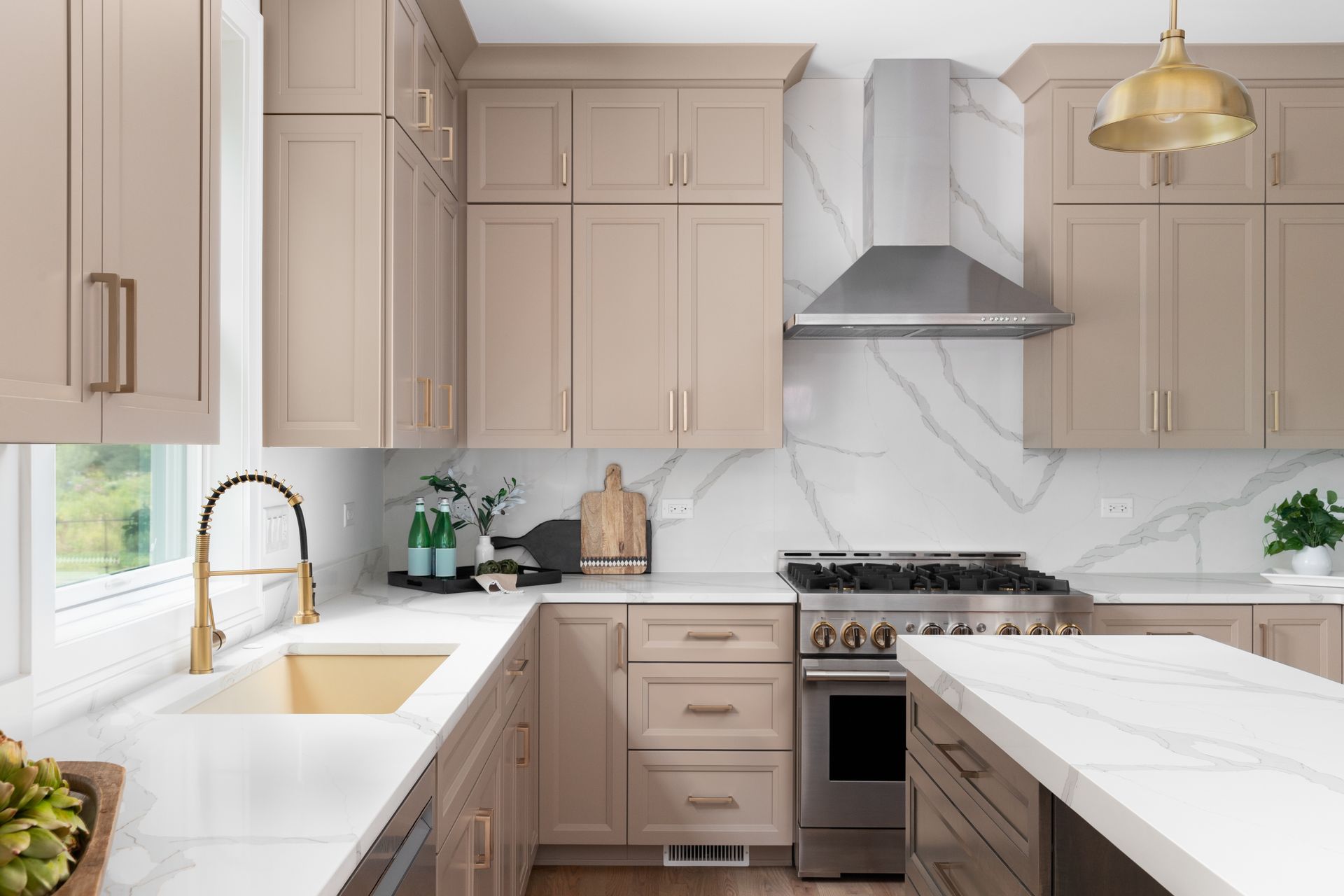 Modern beige kitchen with marble counters, gold pendant light, and stainless steel range hood