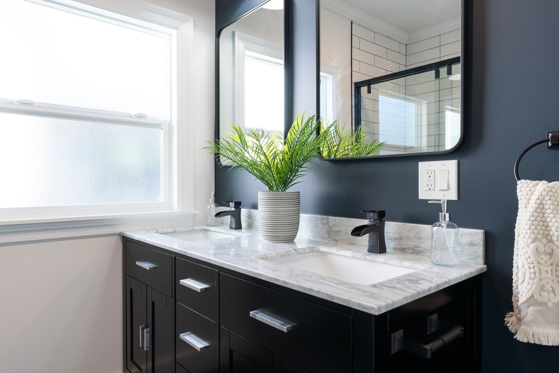 Modern bathroom vanity with black cabinets, white countertop, mirror, and a plant by the window