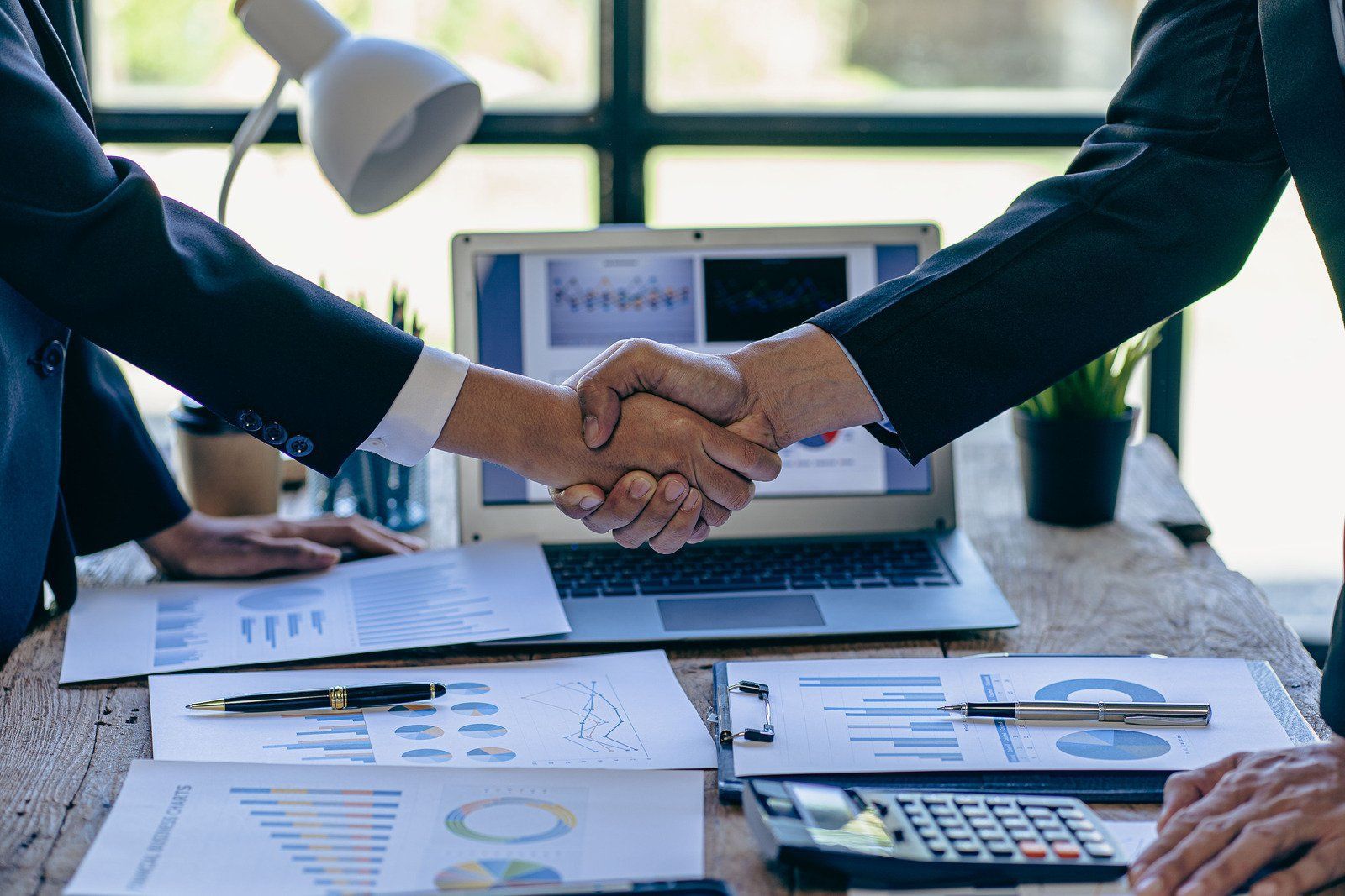 Two businesspeople shaking hands over a desk with financial documents and a laptop, symbolizing a deal.