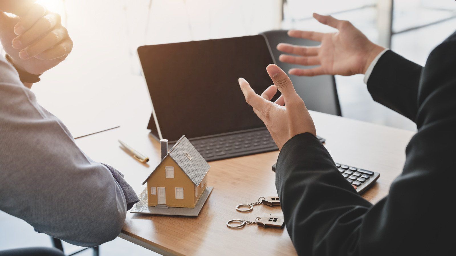 Two people in suits gesturing over a small house model, keys, and a laptop on a desk, suggesting a real estate deal.