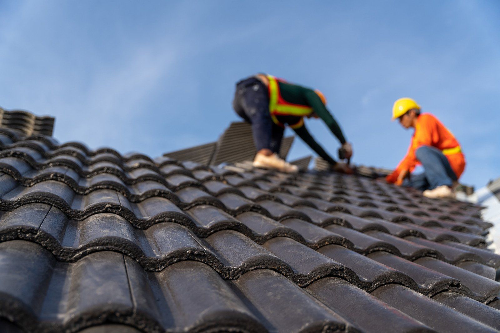 Roofers installing dark tiles on a roof against a blue sky, one in orange, the other in green safety gear.