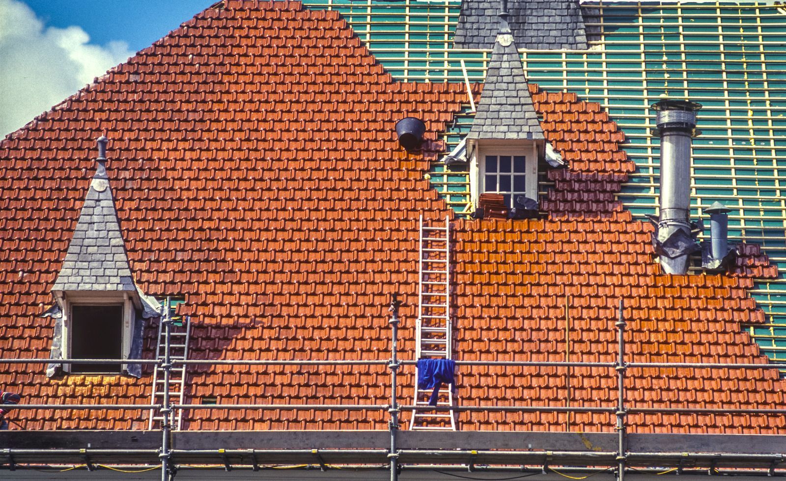 Red tiled roof partially under renovation with scaffolding, ladders, and a chimney against a blue sky.