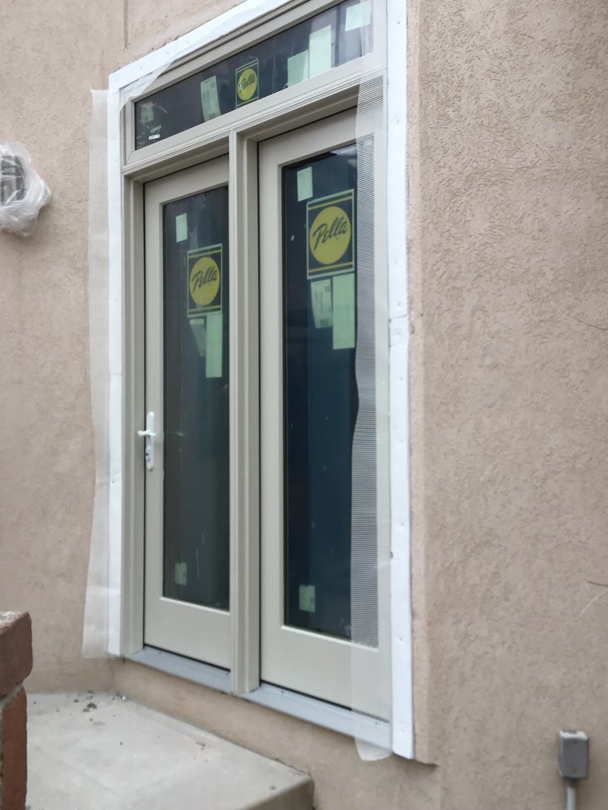 A pair of beige French doors with an upper window, set in a stucco wall. White protective covering surrounds the frame.