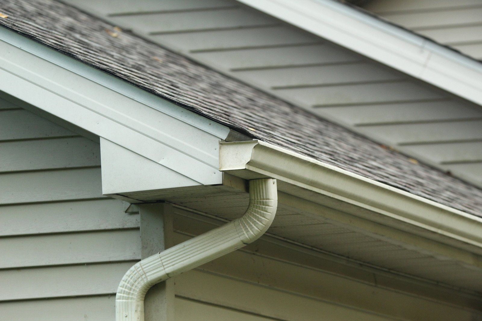Close-up of a house roof with gray shingles, blue gutters, and siding. A white downspout is visible.