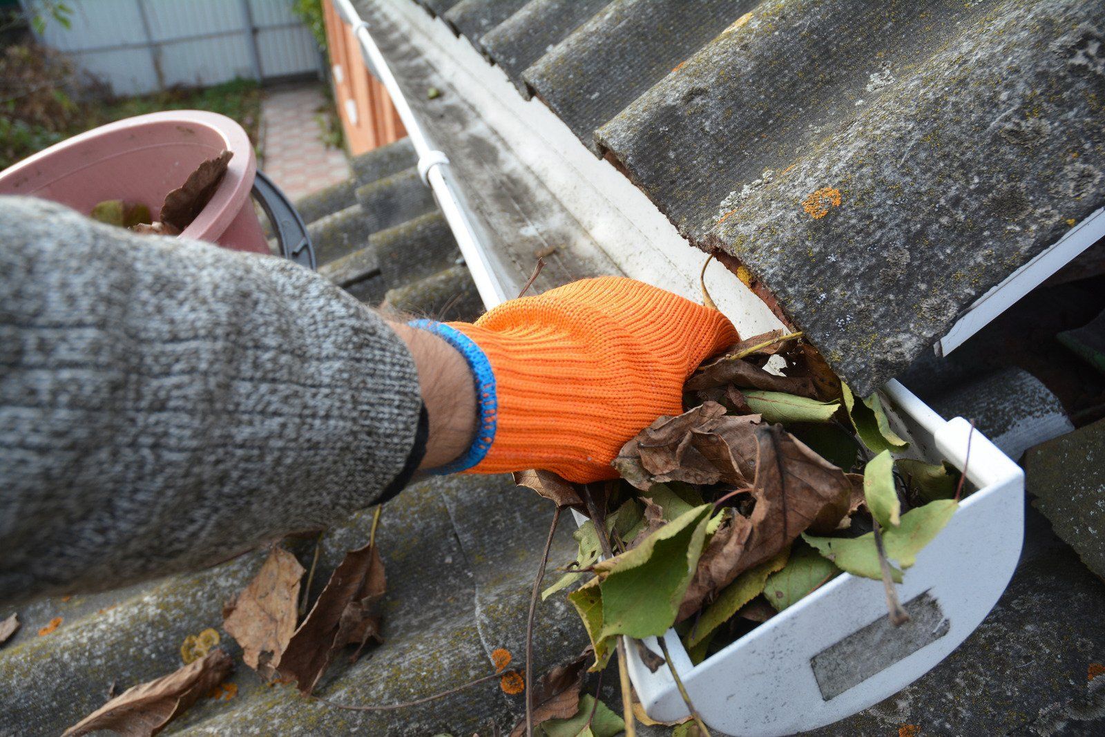 Person wearing orange glove cleaning a roof gutter filled with leaves. A bucket sits nearby.