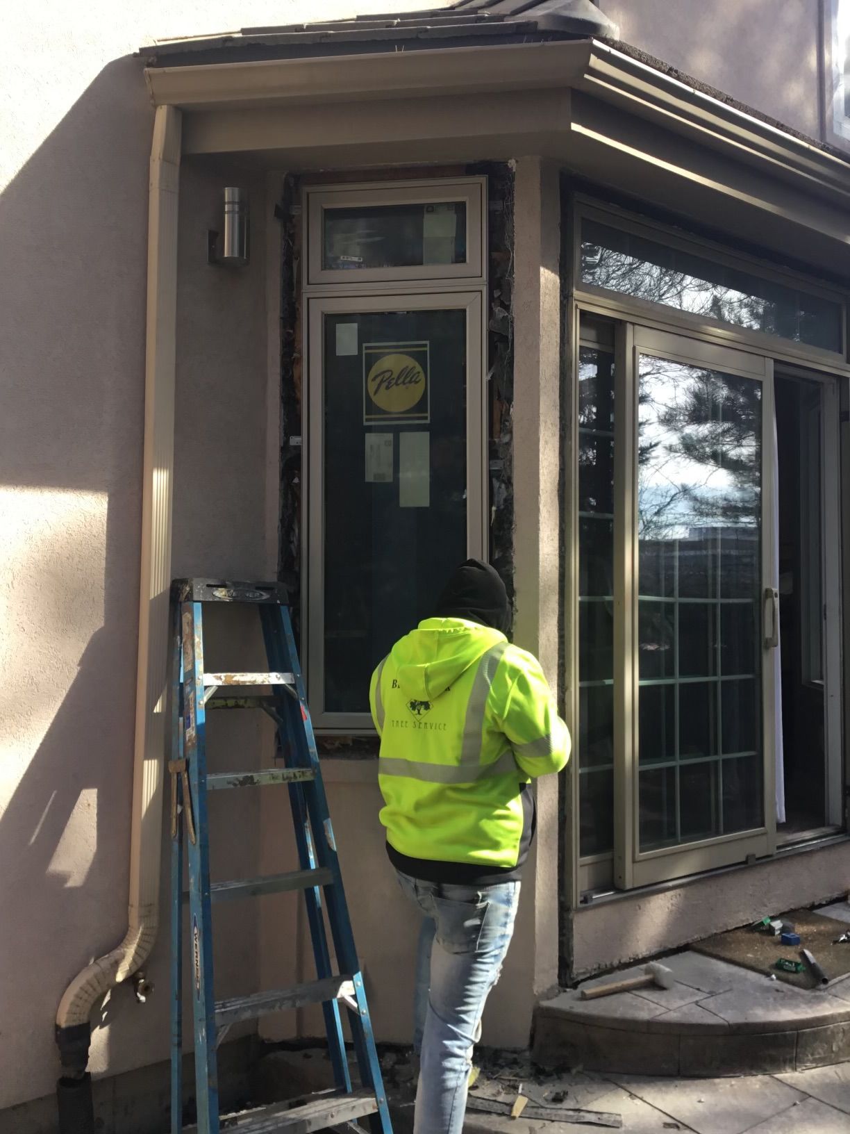A construction worker in a yellow vest stands near a ladder, working on a window installation on a building's exterior.