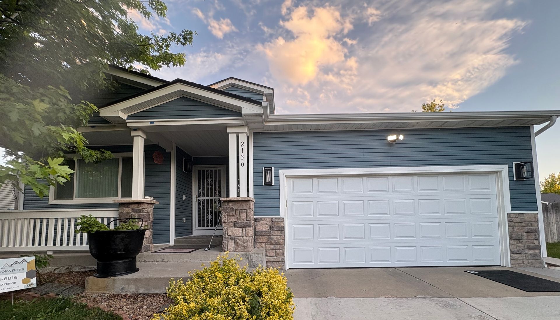 Blue house with a white garage door and porch, set against a partly cloudy sky. A front yard with plants is visible.