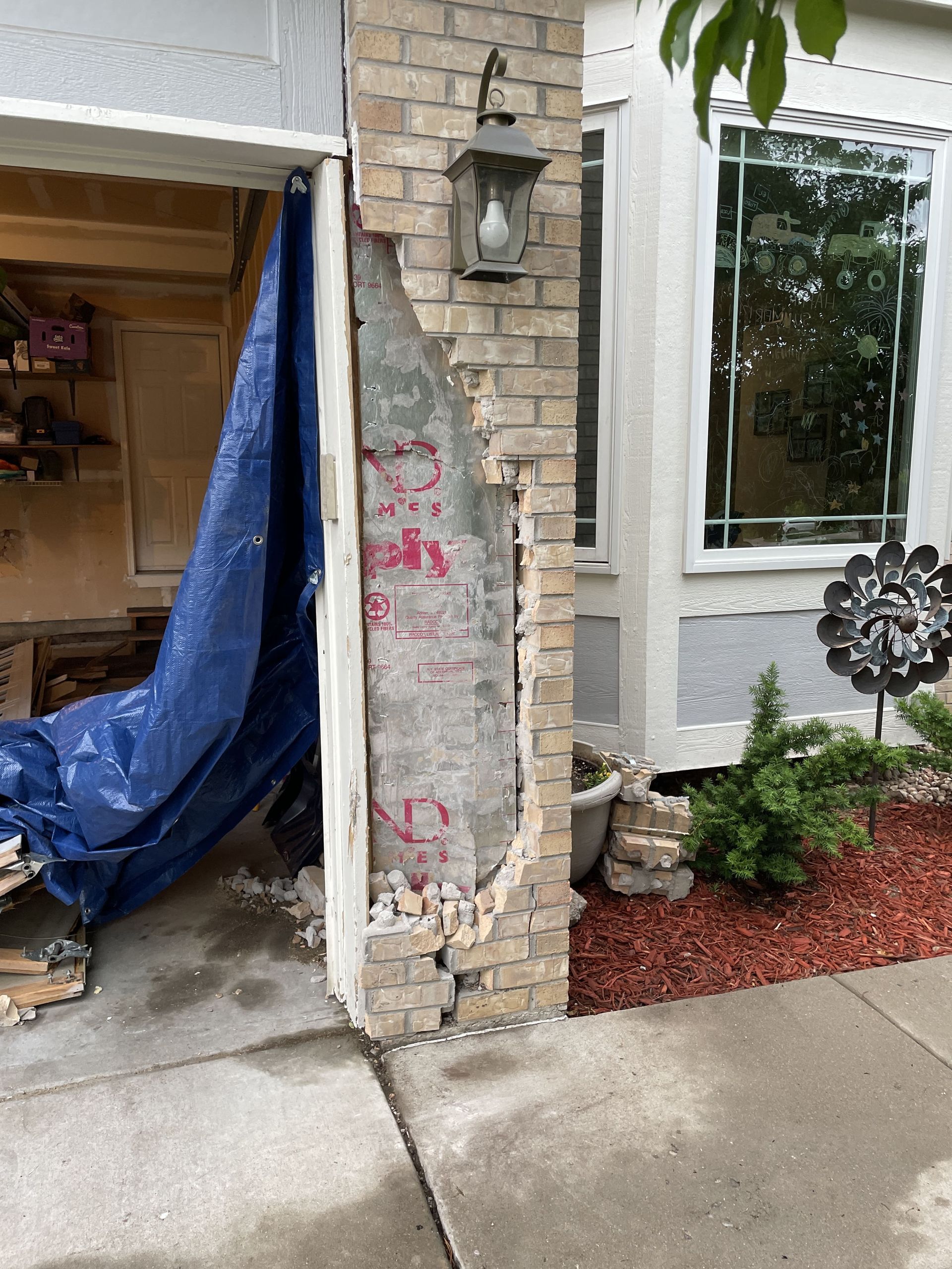 Damaged brick pillar next to a garage door. A blue tarp covers the damaged area. A window and garden are on the right.