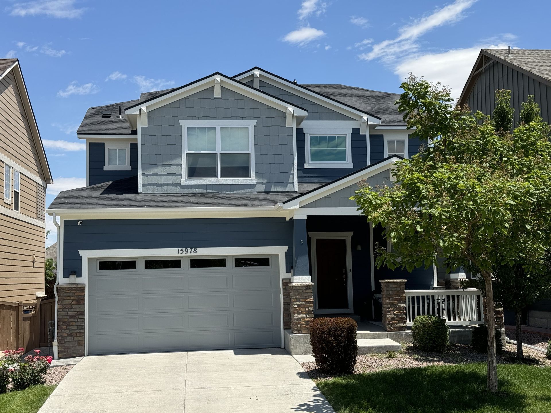 Blue two-story house with a gray garage door, a small porch, and a tree in the front yard under a blue sky.