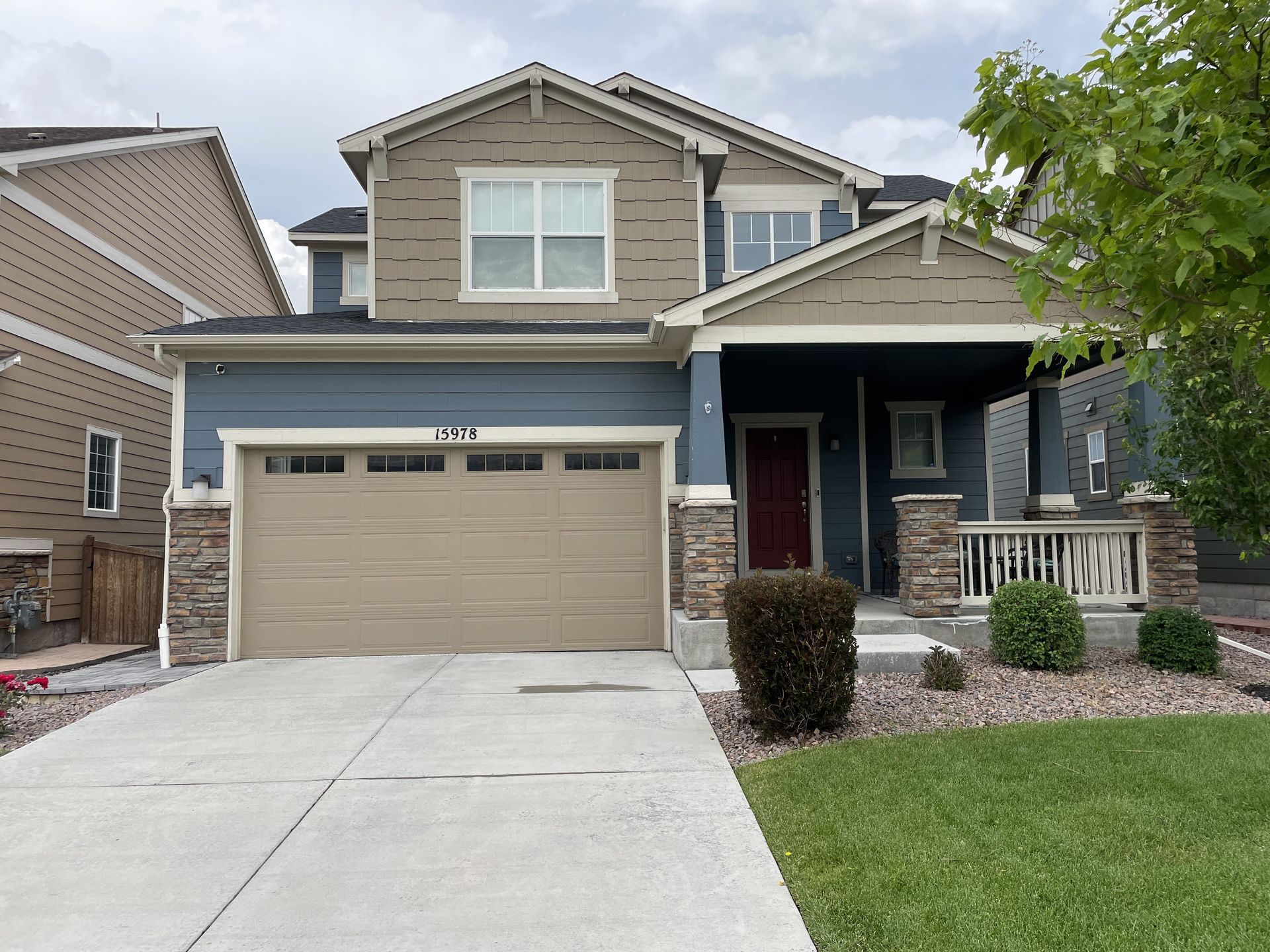 Two-story house with blue and tan siding, a tan garage door, and a concrete driveway. Front porch has stone accents.