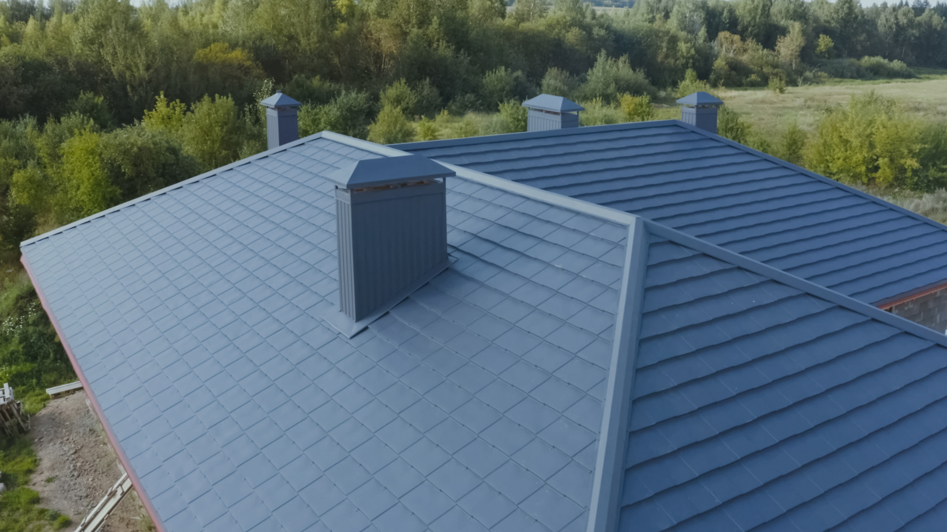 Blue metal roof of a house with three chimneys, set against a backdrop of green trees.