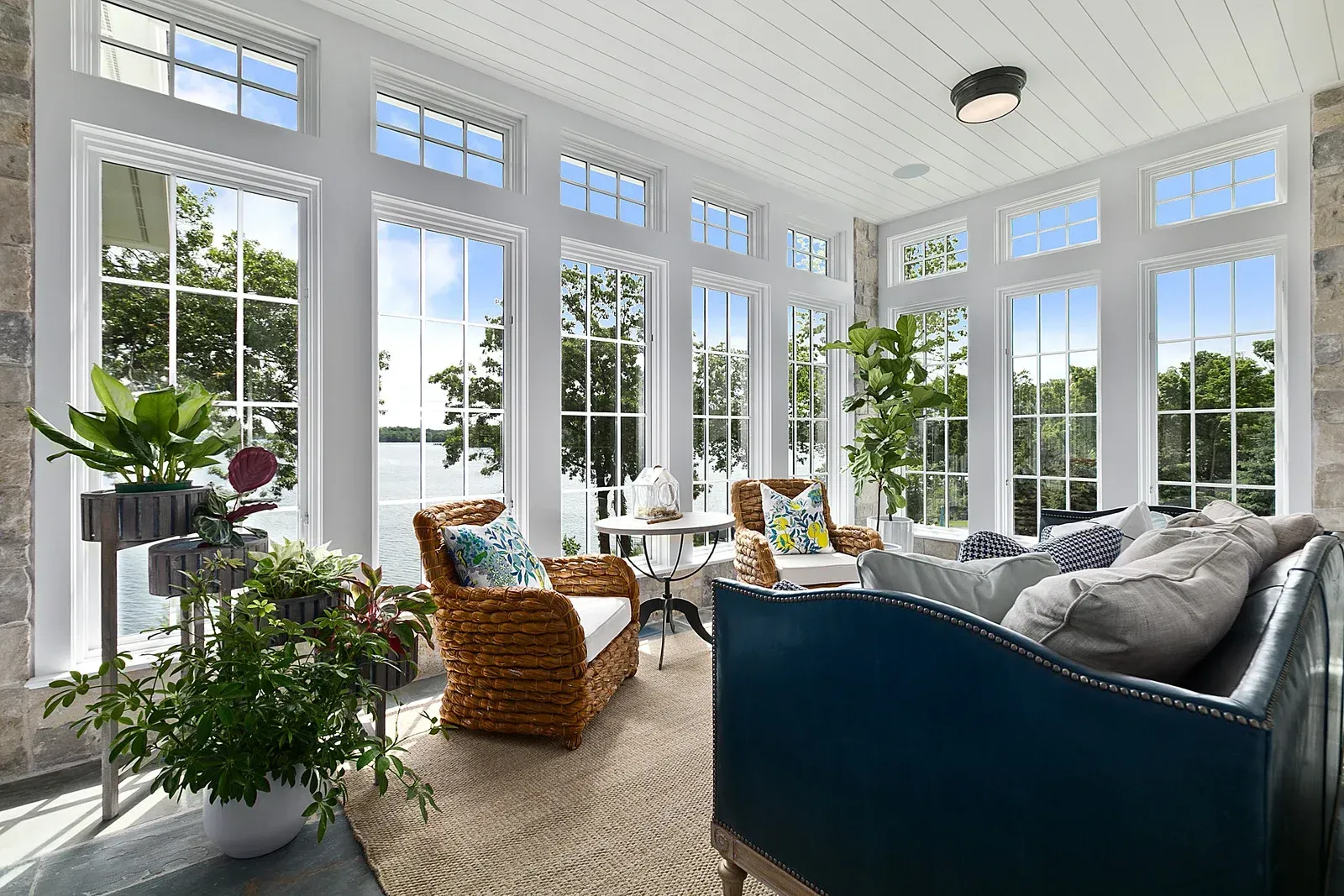 Sunroom with large windows overlooking water, furnished with seating and plants.
