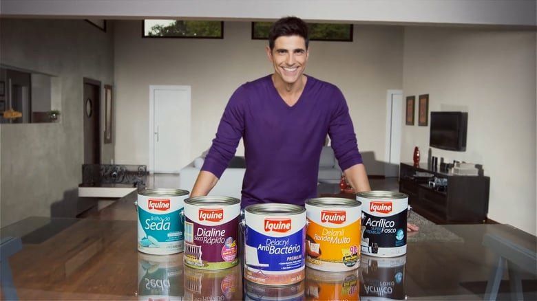 Um homem de camisa roxa está parado em frente a uma mesa com latas de tinta.