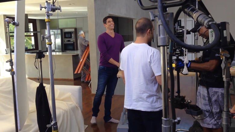 Um homem de camisa roxa está em uma sala conversando com outro homem de camisa branca.