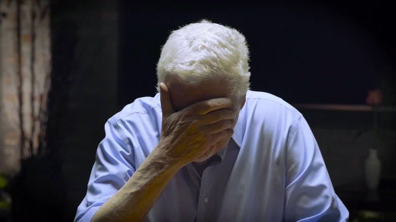 Um homem mais velho cobre o rosto com a mão em um quarto escuro.