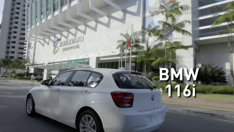 Um bmw 116i branco está dirigindo por uma rua em frente a um prédio.
