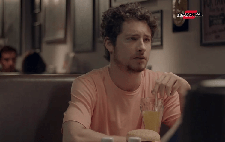 Um homem de camisa rosa está sentado à mesa com um copo de suco de laranja