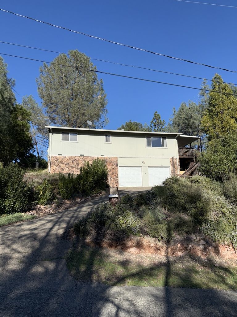 Two-story beige house with a brick facade on a hillside. Two-car garage, deck, and trees. Clear blue sky.