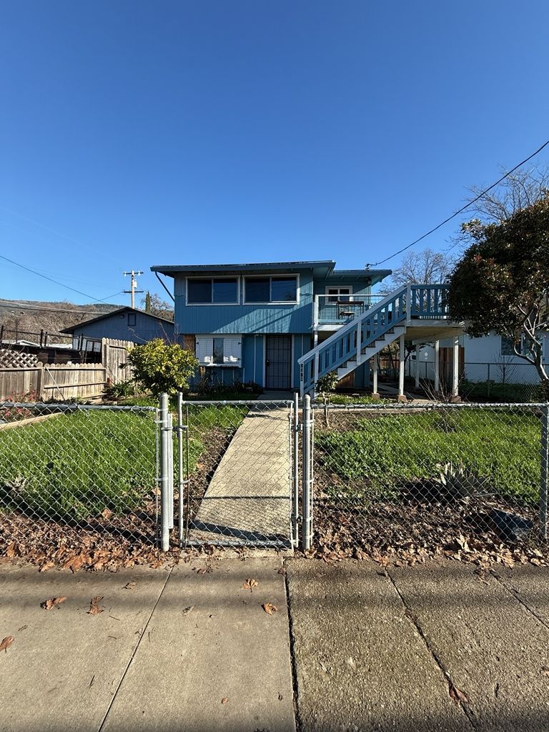 Blue two-story house with a wooden staircase and a chain-link fence on a sunny day.