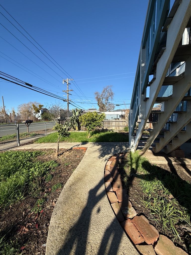 Concrete walkway beside a house with stairs. Green grass and clear blue sky overhead. Power lines in view.