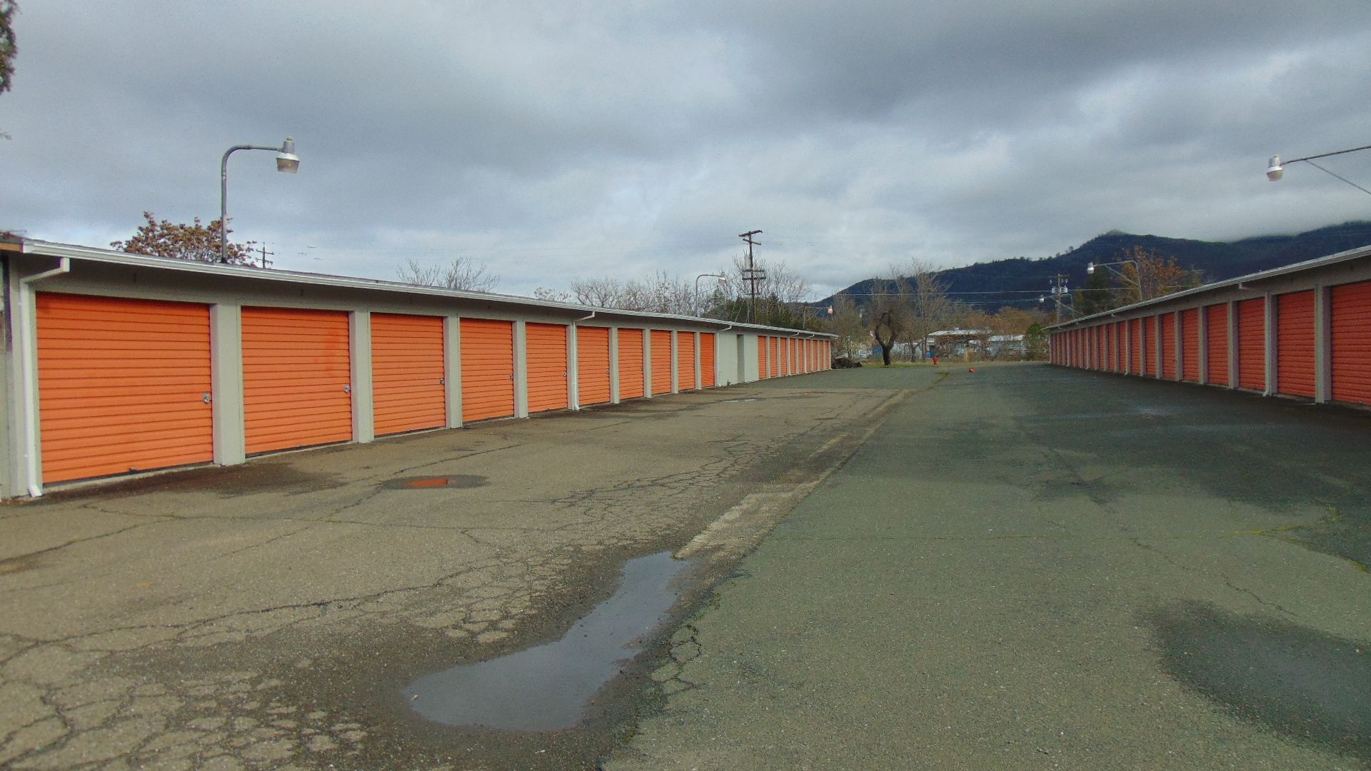 Rows of orange storage unit doors along asphalt road, under cloudy sky.
