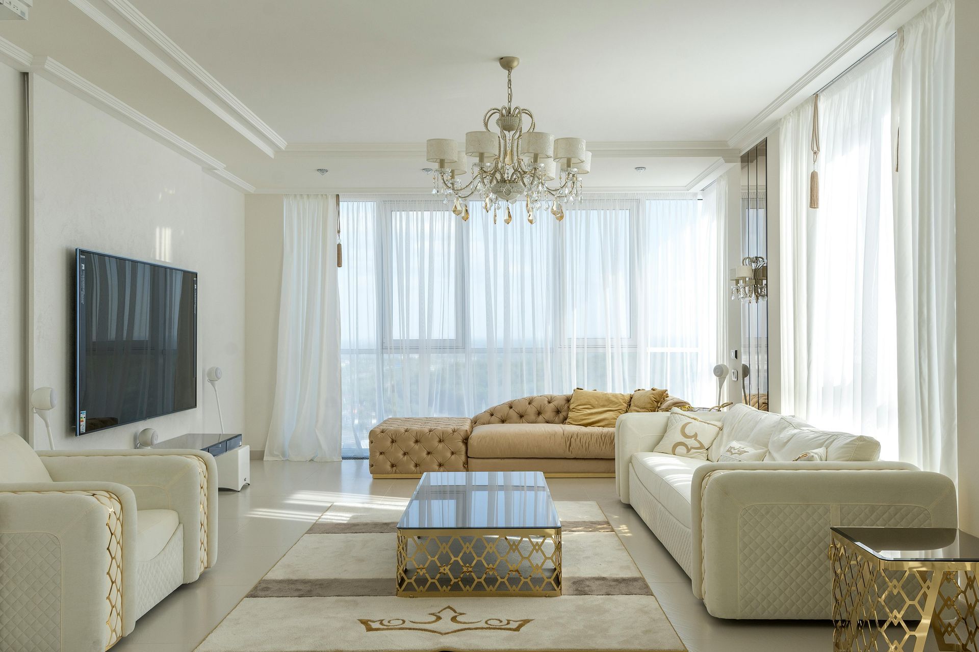 Elegant white living room with gold accents, large windows, and a crystal chandelier.