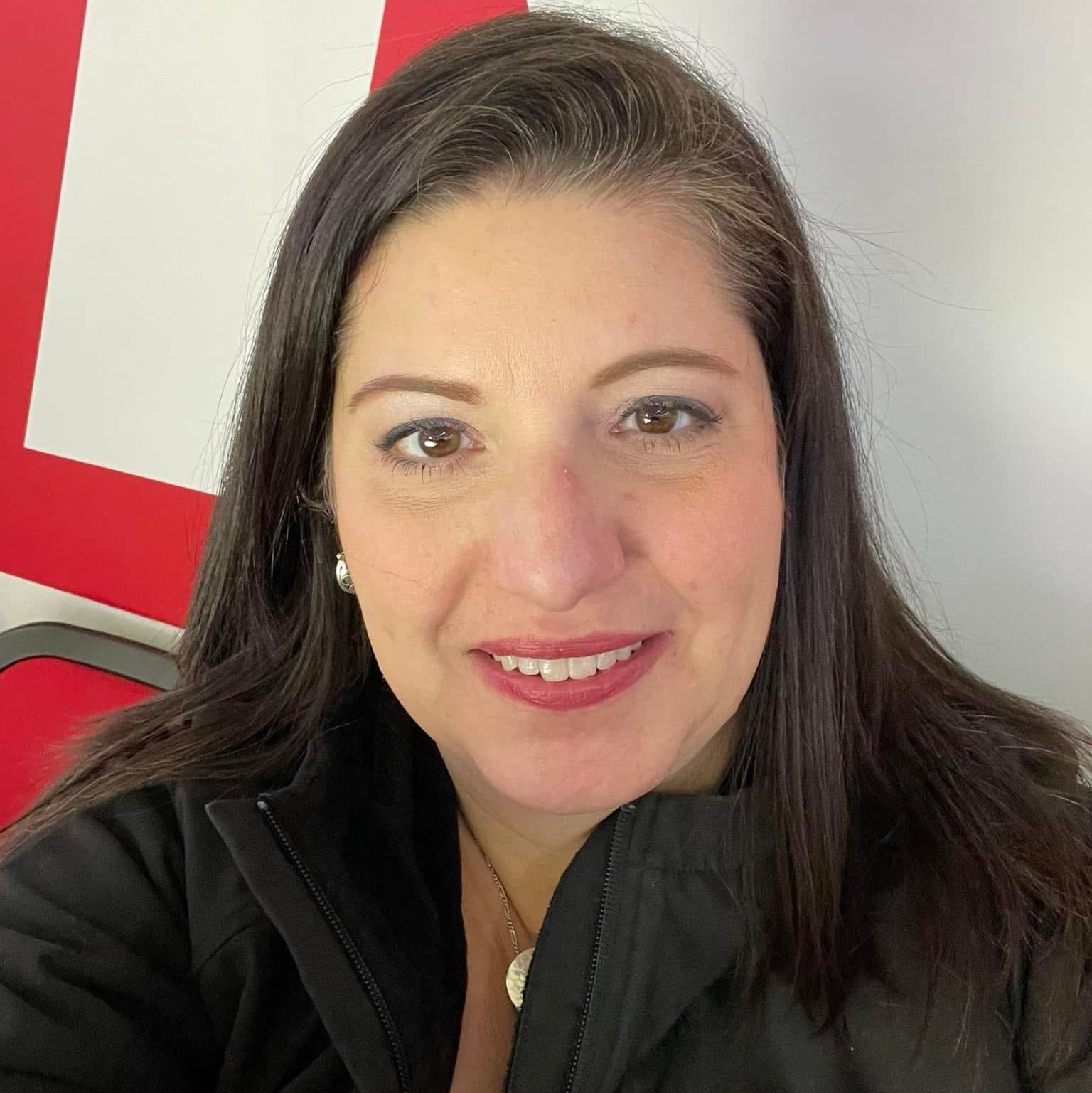 Woman with dark hair smiles at the camera, wearing a black jacket, in front of a red and white background.