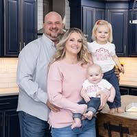 Family in kitchen. Woman holding baby, man, and girl standing. Light skin tones.