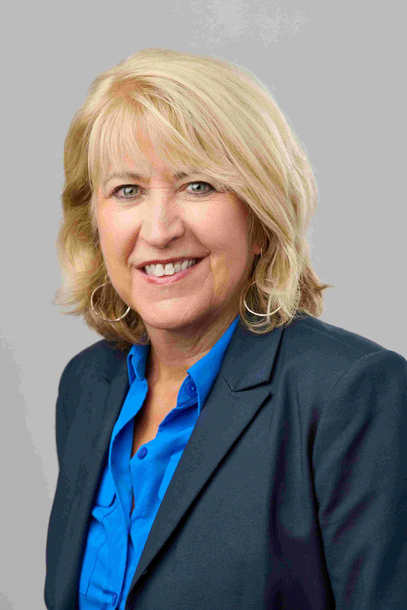 Woman with blonde hair wearing a blue shirt and navy blazer smiles against a gray background.