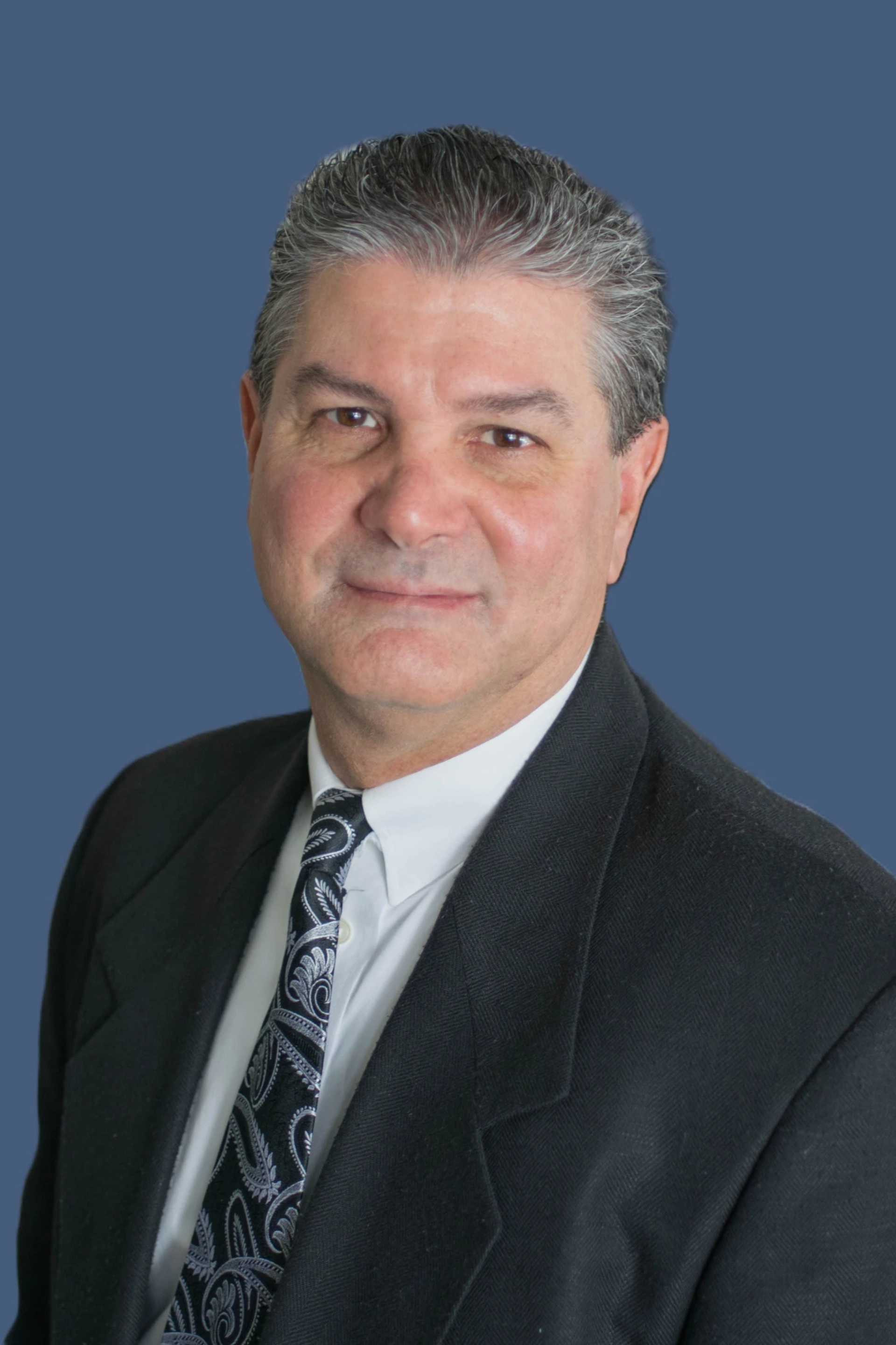 Man in suit, smiling, with dark hair, against a blue background.