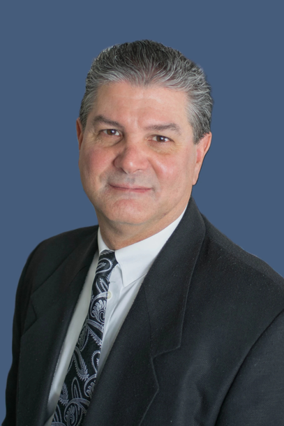 Man in suit, smiling, with dark hair, against a blue background.