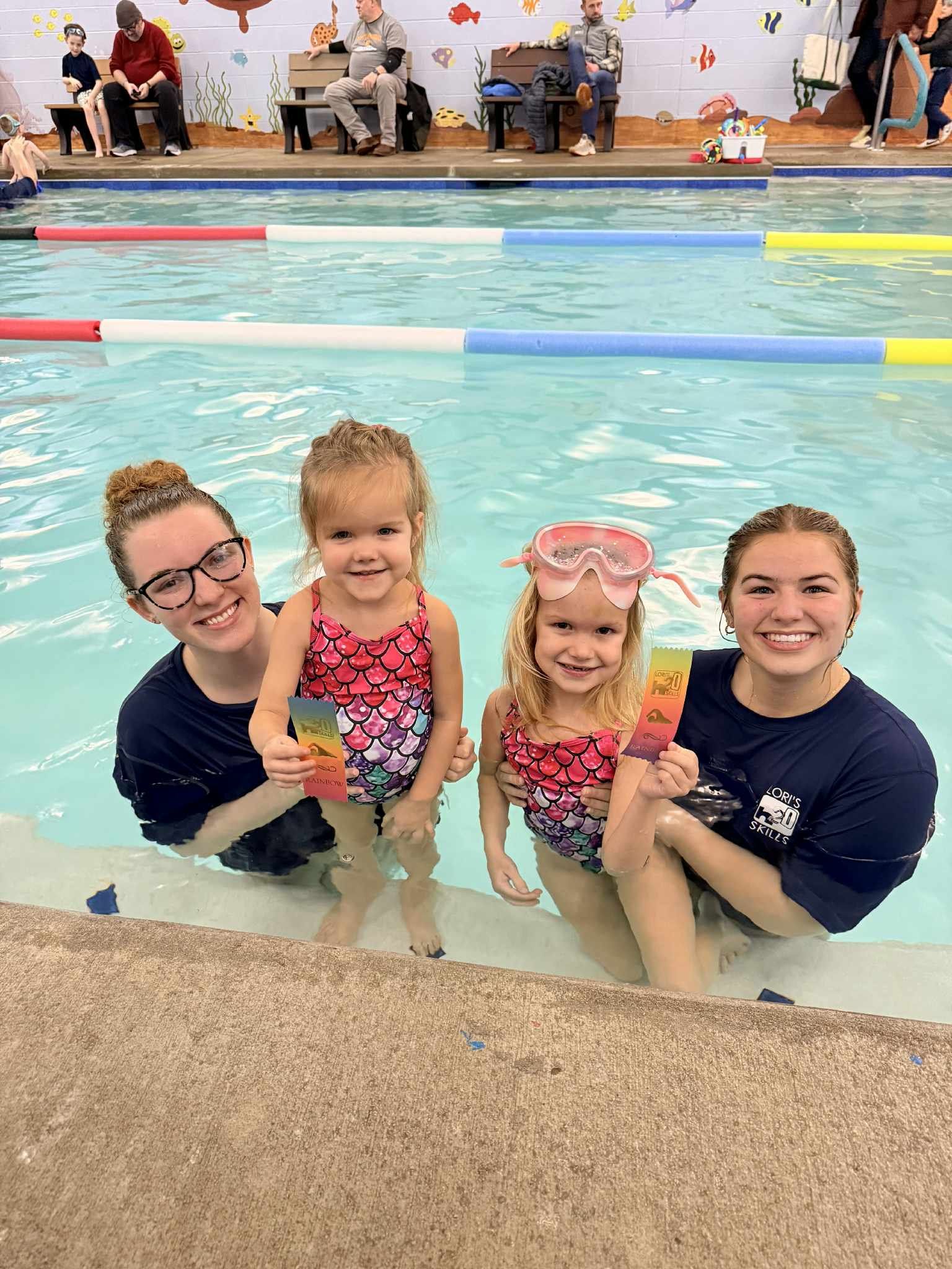 Two adults with two children in a swimming pool. They hold colorful toys, smiling. Other people sit in the background.