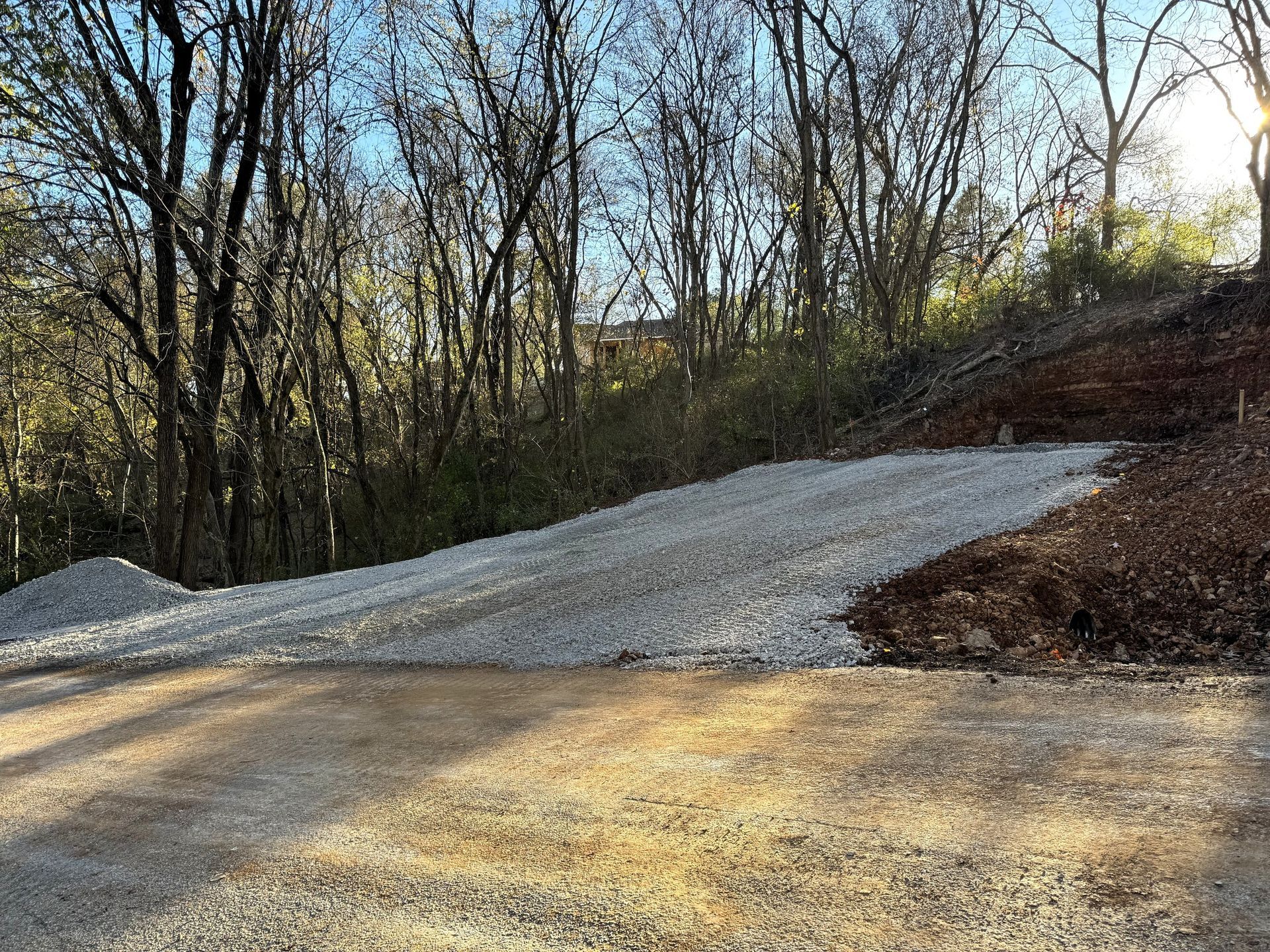 Gravel and dirt driveway leading uphill towards trees.  Sunlight illuminates the scene.